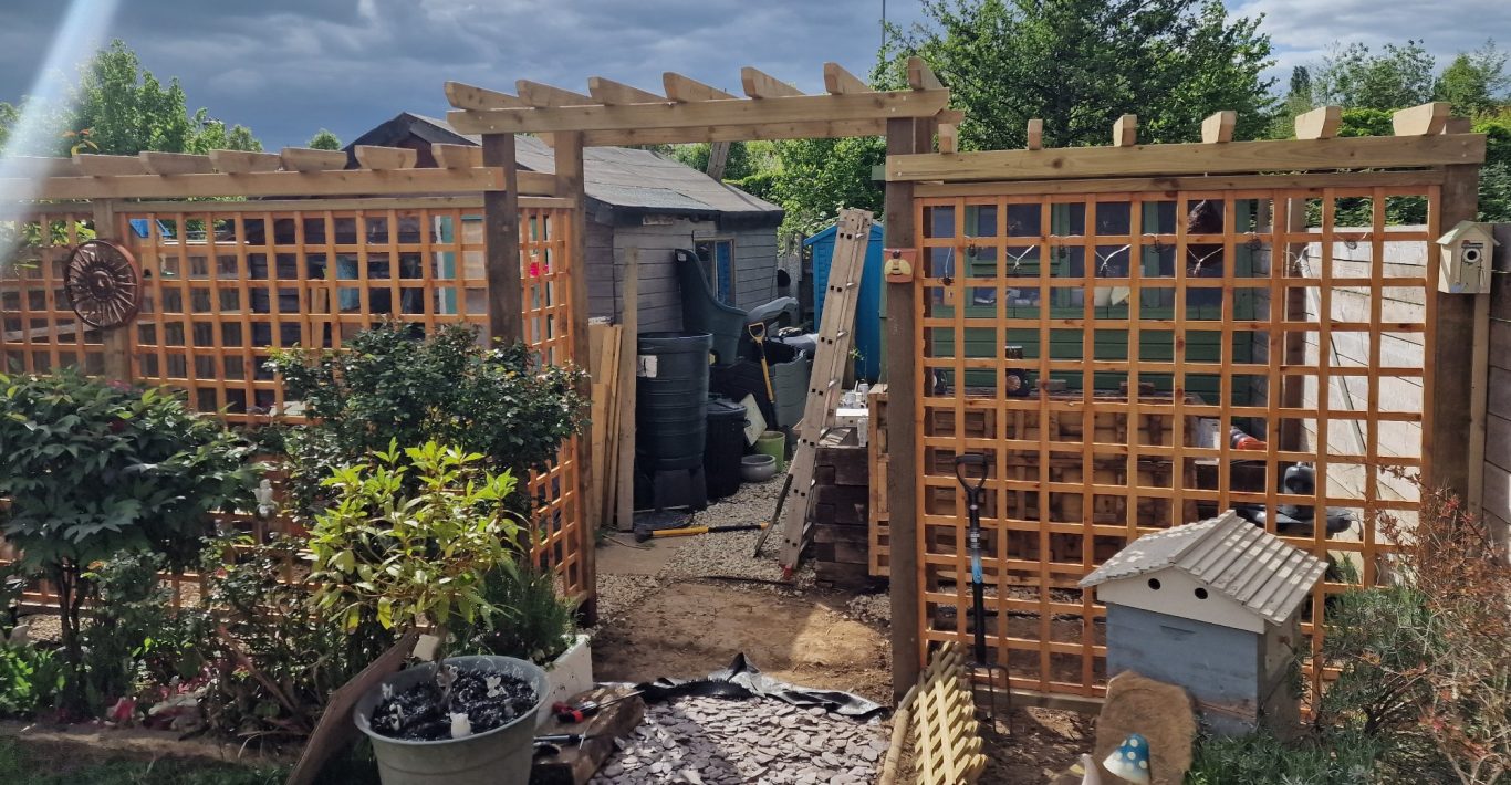 Wooden lattice gate leading to a garden with various plants and gardening tools visible.