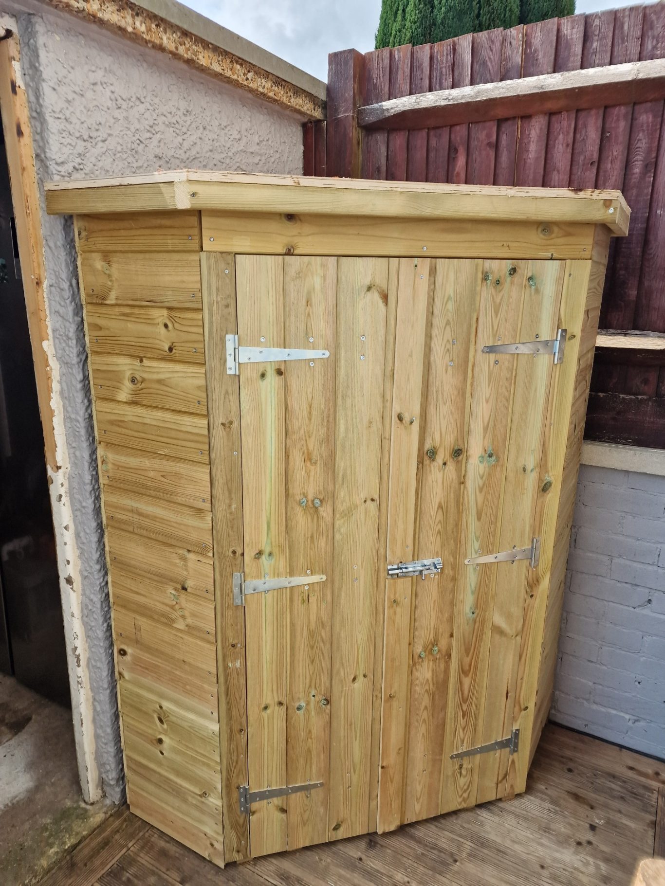 Wooden storage shed with hinged doors, placed against a fence.