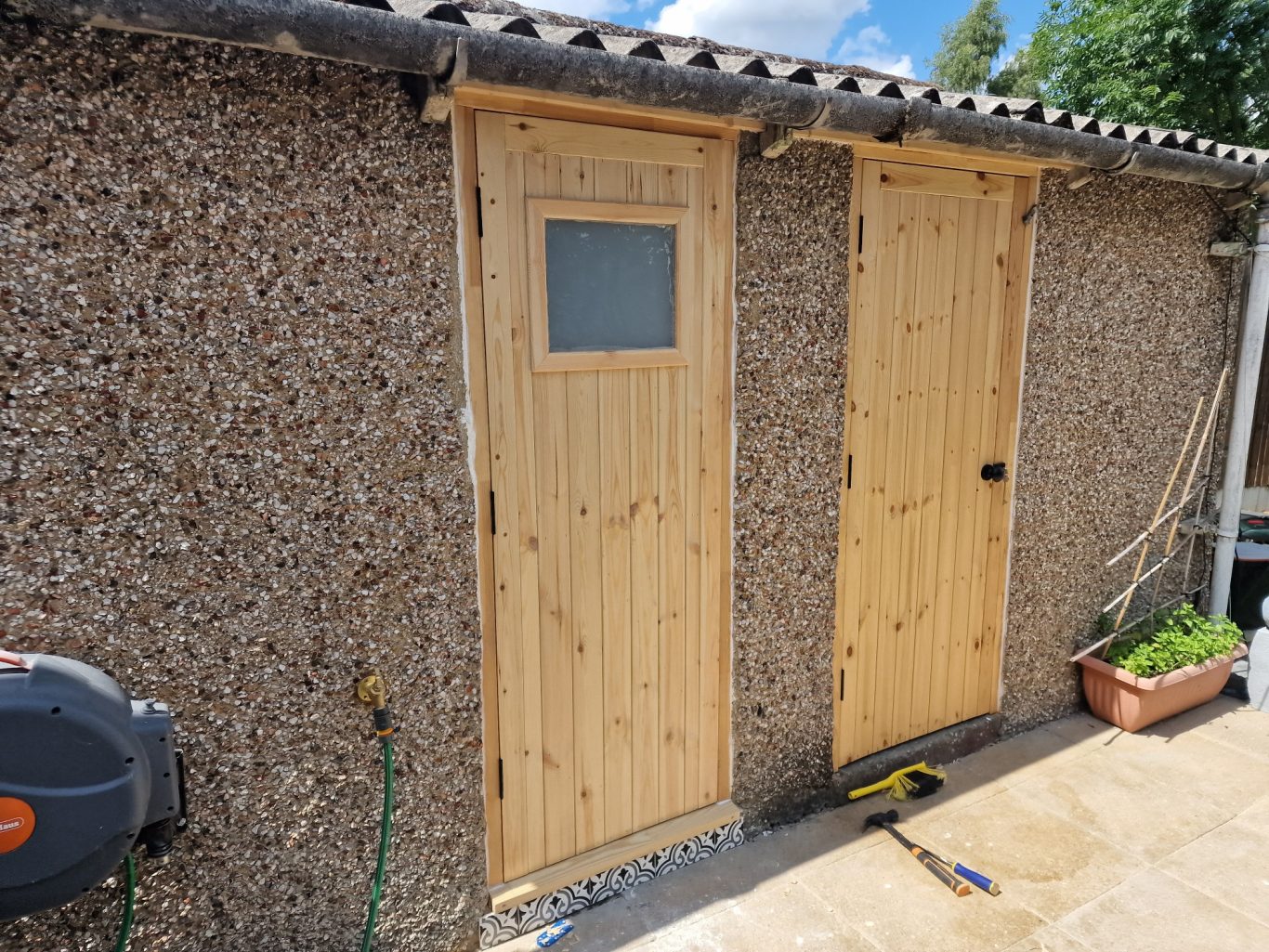 Two wooden doors on a pebble-dashed wall, one with a window, and a plant nearby.